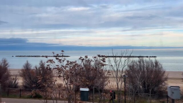 Seaside coastline at sunset seen through the window of a moving train. Reflections on the glass. Waves, breakwater rocks, and tourist facilities such as bars and resorts pass by in the foreground