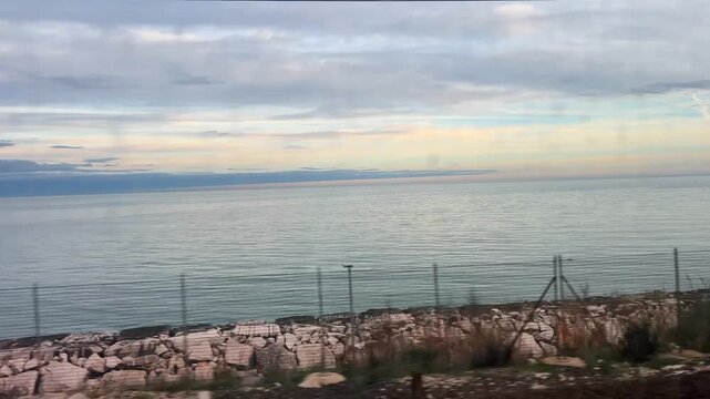 Seaside coastline at sunset seen through the window of a moving train. Reflections on the glass. Waves, breakwater rocks, and tourist facilities such as bars and resorts pass by in the foreground