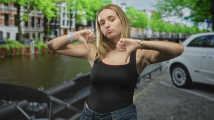 Obraz premium Woman wearing jeans and tanktop showing thumbs down gesture and pouting on a street canal beside a parked car; disapproval rejection attitude.