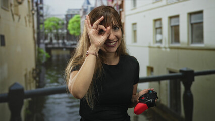 Woman making ok sign with hand while holding red gamepad on building railing overlooking canal  playful gaming. © Krakenimages.com
