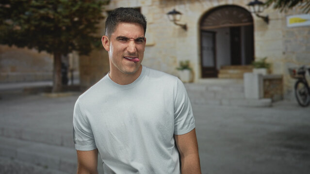 Man sticks tongue out while tilting his head and smiling slightly in front of a stone building entrance on a sunny street; playful curiosity.