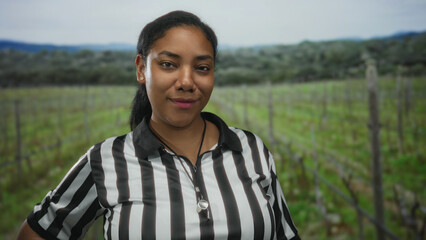 Fototapeta premium Referee woman wears black and white striped shirt and stands smiling with whistle in forest among vineyard rows and rolling hills; serenity.