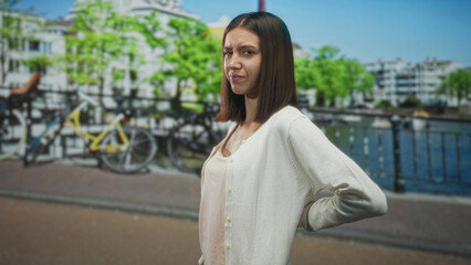 Fototapeta premium Woman standing with hand on lower back for backache, wincing beside parked bicycles along a canal side railing on a street; discomfort concern.