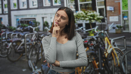 Naklejka premium Young woman with hand on chin and folded arm, finger touching jaw in a street with parked bicycles and metal racks; contemplation.