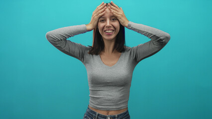 Young woman with hands on head and open mouth, wearing gray long sleeve top and jeans with visible navel against a turquoise studio wall; surprise expressive pose.