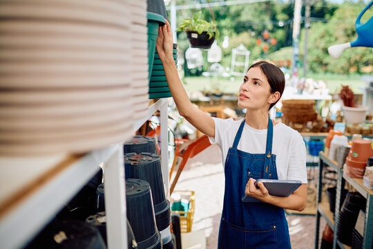 Woman wearing apron using a digital tablet for inventory management at a garden center, checking empty flower pots on shelves