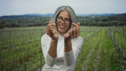 Fototapeta premium Middle aged woman makes finger heart gesture with hands in a green field during daytime; joy connection.
