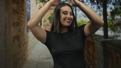 Fototapeta premium Woman smiling with hands above head revealing bare arms on a narrow street near stone buildings; joy casual movement.