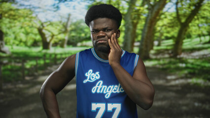 Man with hand on cheek for toothache in forest wearing blue basketball jersey and number 77,...