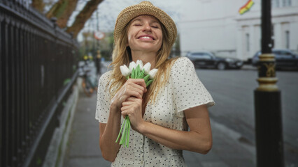 Fototapeta premium Woman wearing straw hat holds white tulips to chest on street with eyes closed and smiling while hugging bouquet; serenity spring memory.
