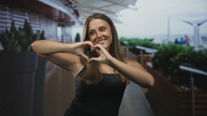 Fototapeta premium Young woman in black tank top forms heart with hands, smiling near large planters and railing on an airport terrace under canopy; warm affection.