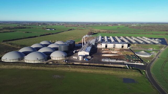 Danish biogas facility featuring large digesters, storage tanks, and processing buildings surrounded by agricultural fields. The scene highlights modern renewable energy production and sustainable was