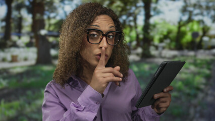 Woman in purple shirt and glasses holding tablet with finger to lips for silence in leafy park under sunlight  privacy. © Krakenimages.com