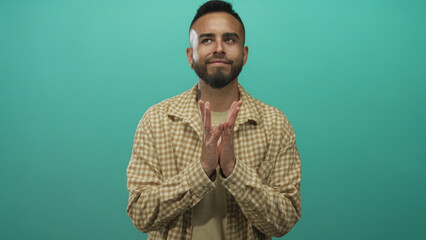 Obraz premium Young hispanic man with hands pressed together and a slight smile wearing a checked shirt in a turquoise studio; calm gratitude.