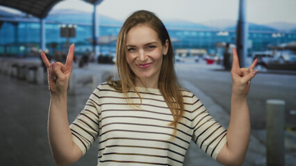 Fototapeta premium Young smiling caucasian woman in striped shirt raises both hands in rock horns gesture at airport terminal; excitement.