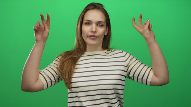 Young caucasian woman wearing striped top raises both hands to form air quotes gesture in green studio; sarcasm.
