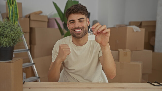 Man holds keychain and raises fist inside residential building surrounded by cardboard boxes and potted plants; ownership pride.
