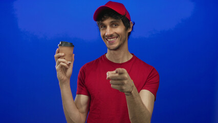 Naklejka premium Man wearing red cap and t shirt holds ribbed coffee cup and points finger toward viewer in vivid blue studio; invitation.