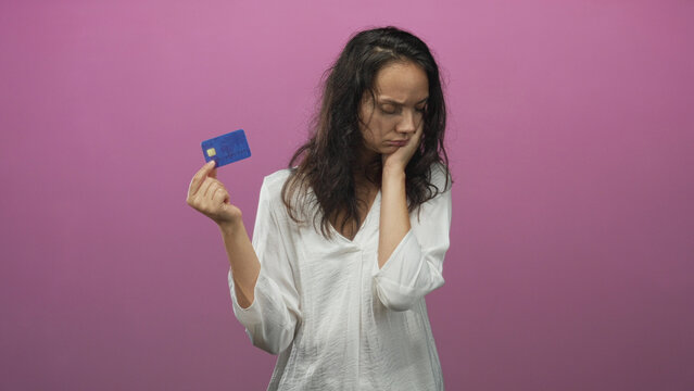 Woman holding blue creditcard with left hand and hand on cheek in pink studio, showing worried expression and slouched posture; financial worry.