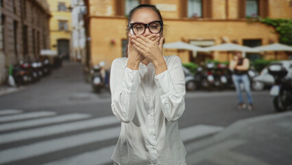 Fototapeta premium Woman covering her mouth with hands and making a shushing gesture on a city street near scooters and a cafe; surprise silence.