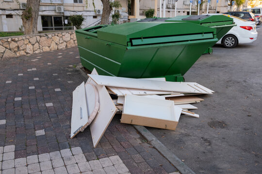 Broken white wooden furniture piled next to large green waste dumpsters