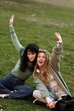 Friends enjoying a sunny day outdoors, smiling and waving while sitting on green grass in a park setting, capturing joyful moments together during afternoon
