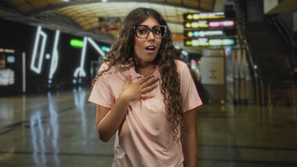 Fototapeta premium Hispanic woman touching hand to chest in airport terminal, wearing glasses and pink shirt, mouth open; surprise travel moment.