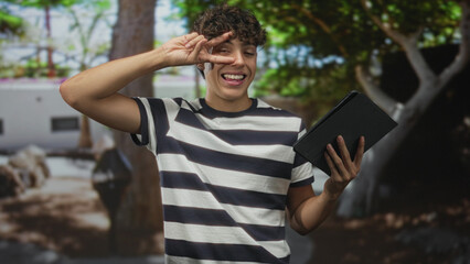 Man holding a tablet makes a peace sign over his eye while standing near a building wearing a striped shirt and smiling  playful youth. © Krakenimages.com