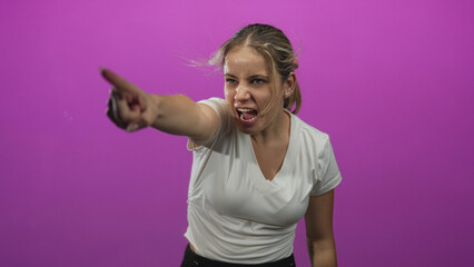 Fototapeta premium Young blonde woman points finger in magenta studio with arm extended and clenched jaw and firm stance; frustration.