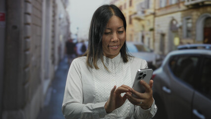 Fototapeta premium Woman tapping smartphone screen with both hands on a narrow city street beside parked car and old buildings; serenity.