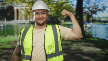 Fototapeta premium Man engineer in hardhat and high visibility vest smiles and flexes arm beside park building and pond; confidence.
