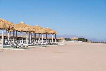 Fototapeta premium Beachfront view of thatched-roof cabanas and lounge chairs on sandy shore under clear blue sky with distant mountains visible