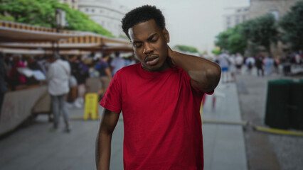 Fototapeta premium Young american man wearing red tshirt touching neck in street market amid blurred stalls;tension strain fatigue.
