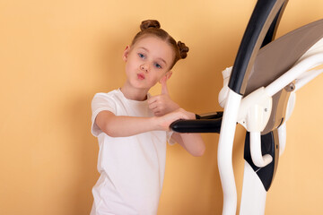 Smiling young girl in white t-shirt giving thumbs up while standing on fitness machine against a light orange background.