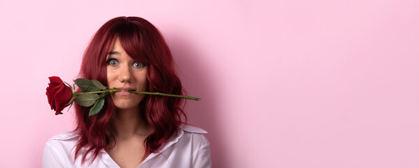 Young woman with red hair holding a red rose in her mouth, smiling playfully against a pastel pink background, wearing a white shirt. panoramic layout
