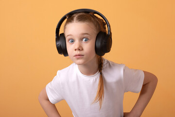 Young girl in white shirt and headphones, wide-eyed and surprised, standing against an orange background. Studio shot, playful and energetic mood.
