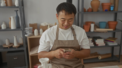 Man taps smartphone screen while seated at ceramic worktable scattered with clay modeling tools in...