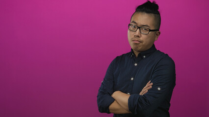 Obraz premium Young man with crossed arms and glasses in studio wearing navy shirt with rolled sleeves against pink backdrop; quiet confidence.