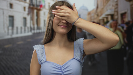 Fototapeta premium Teenage girl in light blue top covers eyes with hand blocking sunlight on bustling city street; shyness.