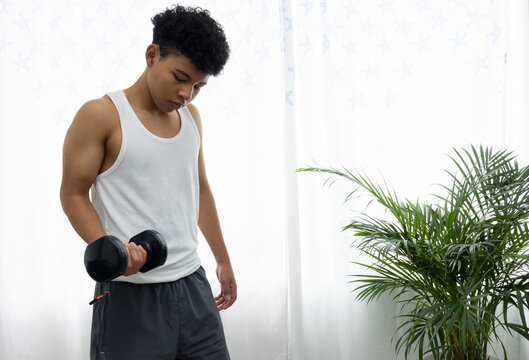 Dark skinned teenager exercising in his living room, lifting a dumbbell with one hand. Home workout, healthy lifestyle, physical activity, and space for advertising text