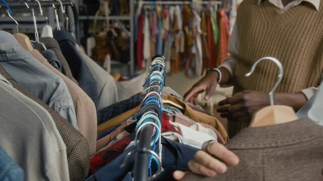 Close-up shot of unrecognizable people going through secondhand clothes on rack in cozy modern thrift shop