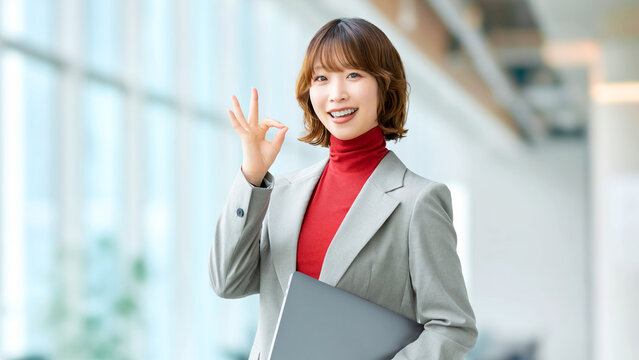 ノートPCを持ちながらOKサインを示す日本人女性社員の前向きなビジネスシーン, Japanese office worker holding a laptop and showing an OK sign, positive business scene