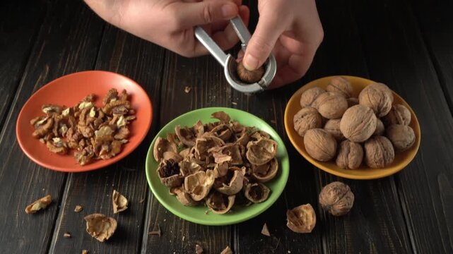 Hands cracking walnuts using a nutcracker, with broken shells and shelled nuts arranged on colorful plates on a wooden table