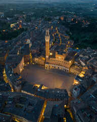 Obraz premium Aerial night view of Torre del Mangia rising above Piazza del Campo in Siena, Tuscany. Illuminated medieval tower and historic city center seen from above during evening hours