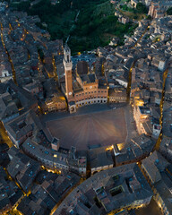 Obraz premium Aerial night view of Torre del Mangia rising above Piazza del Campo in Siena, Tuscany. Illuminated medieval tower and historic city center seen from above during evening hours