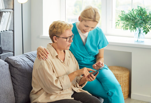 Kind caregiver nurse, senior woman sitting on sofa in home, older patient pointing at smartphone, supportive and attentive young doctor, digital assistance learning during home care or personal helper