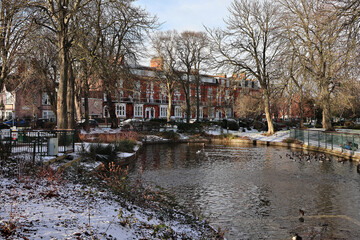 Fototapeta premium Serene snow-surrounded pond and frozen landscape at Tower Gardens, Skegness; winter in picturesque English public park with cold weather and icy duck pond setting in seaside town, Lincolnshire