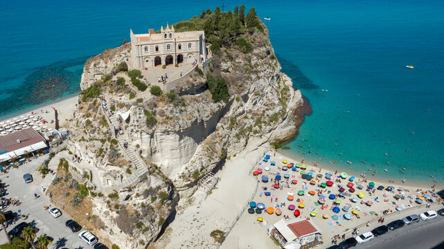 Aerial view of Sanctuary of Santa Maria dell'Isola in Tropea, Calabria, Italy. The ancient church stands on a rocky promontory, overlooking a crowded sandy beach filled with colorful sun umbrellas.