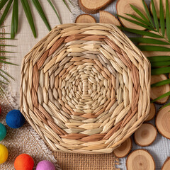 Top view of traditional woven straw plate placed on natural wooden slices with green palm leaves and vibrant colorful wool balls for eco friendly home decor concept