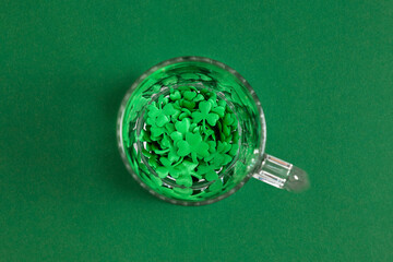 St. Patrick's Day concept: Top view of a glass mug filled with green shamrock confetti on a dark green background. Minimalist holiday background with copy space for Irish celebration.